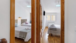 Een smalle gang die uitkomt in een slaapkamer met twee bedden, een televisie en een raam.