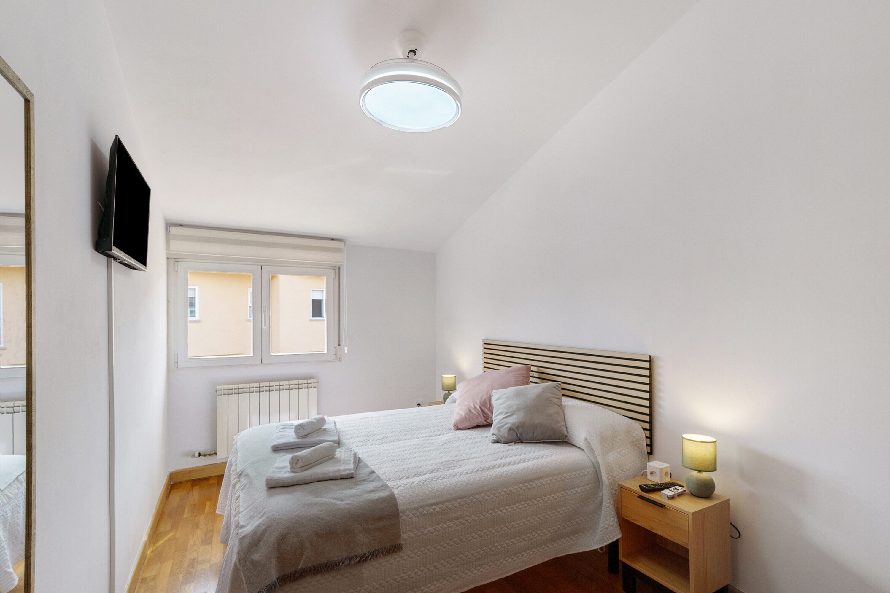 A bedroom with a bed, bedside lamps, a flat-screen TV, a window, and a radiator.