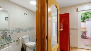Een badkamer met een toilet, een glazen wastafel en een rode deur die uitkomt op een buitenruimte.