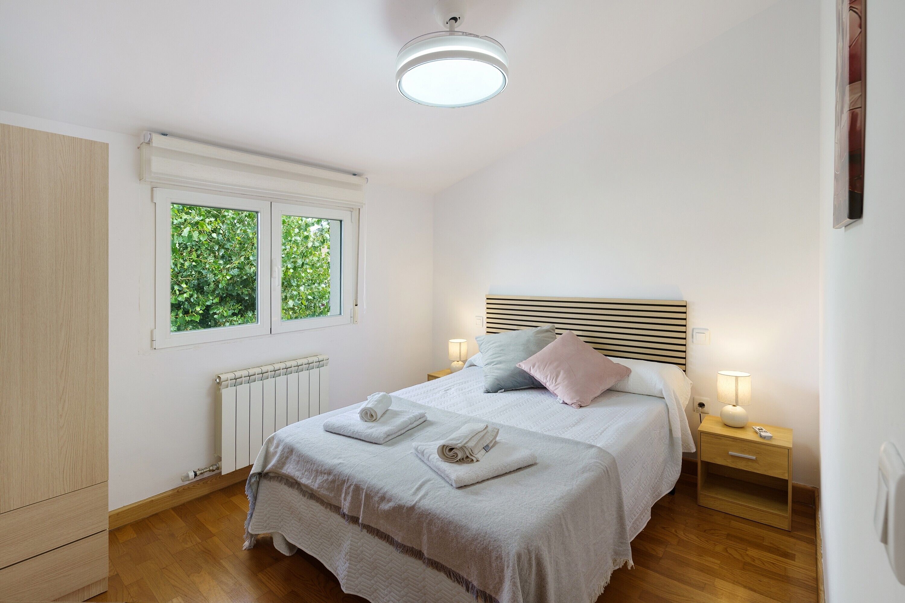 A bedroom with a bed, bedside lamps, a radiator, and a window with greenery outside.