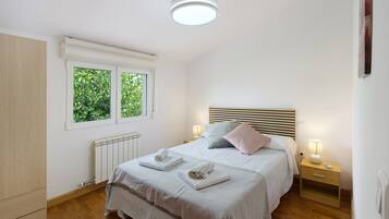 A bedroom with a bed, bedside lamps, a radiator, and a window with greenery outside.