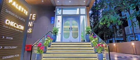 Entrada de hotel con diseño moderno, que incluye una escalera, plantas en macetas y señalización para habitaciones, restaurantes y una piscina.