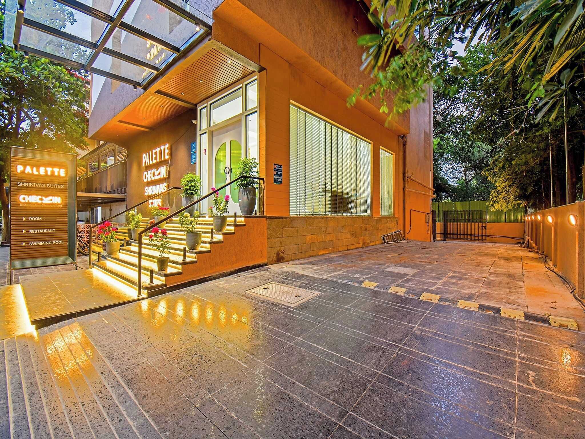 Entrada de un hotel con una escalera que lleva a un segundo piso, un letrero con la leyenda 'PALETTE CHEZIN SHRINIVAS SUITES' y un área de piscina.