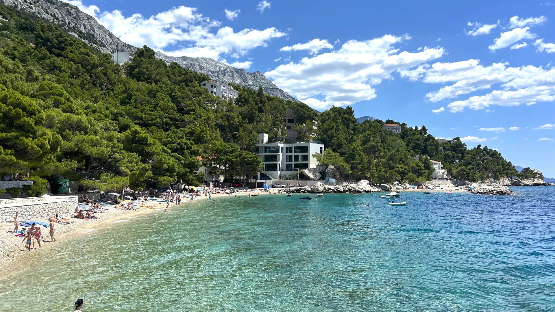 A coastal beach with clear waters, a rocky mountain backdrop, and a building with multiple floors.