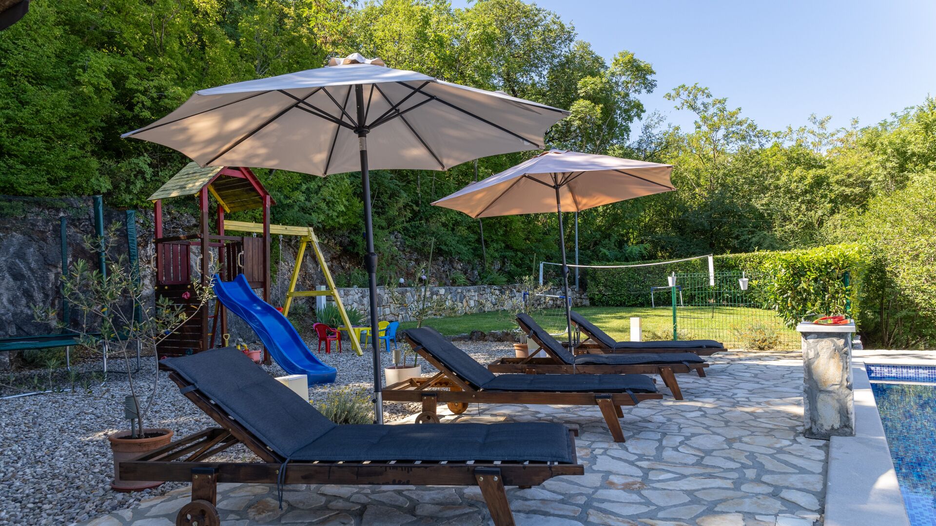 Ein Poolbereich mit Liegestühlen und Sonnenschirmen, einer Kinderspielanlage und einem gepflasterten Weg.