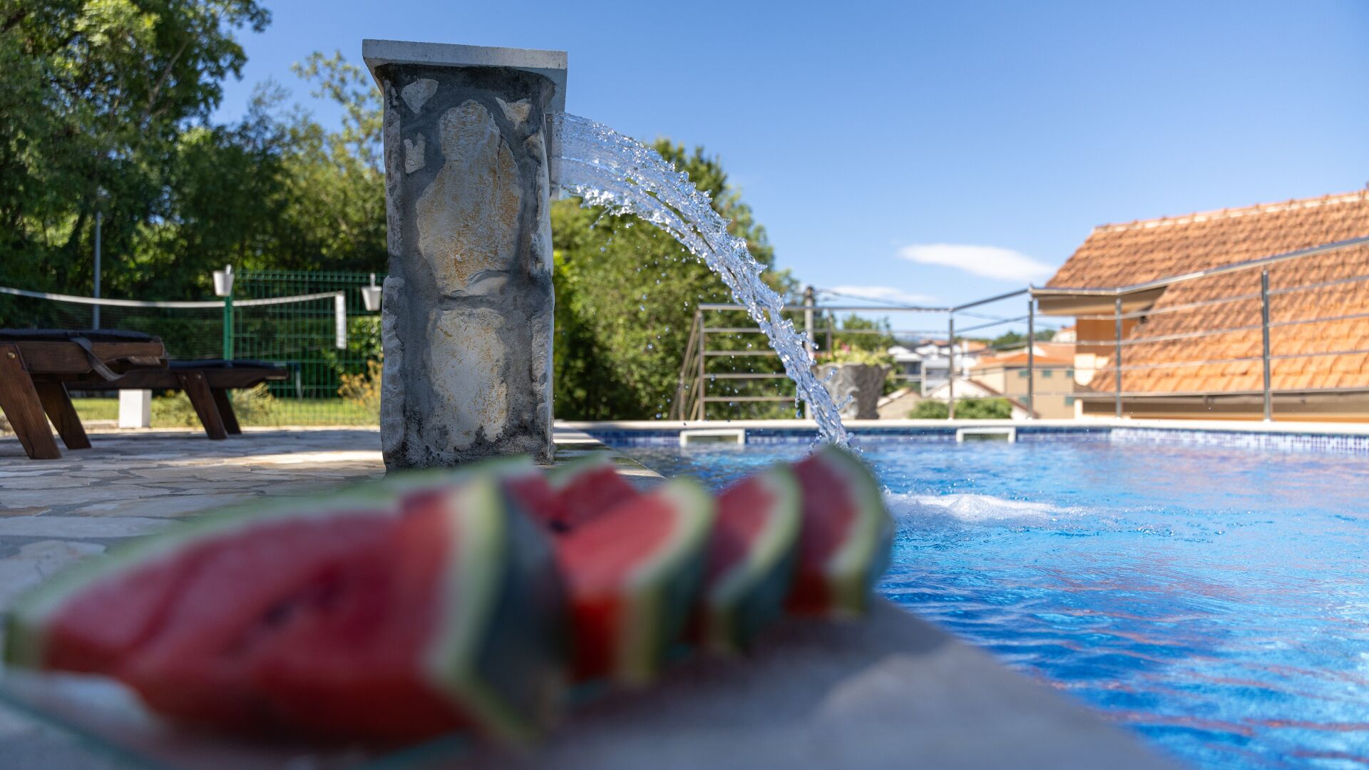 Una piscina con fuente de agua, una rebanada de sandía y un banco.