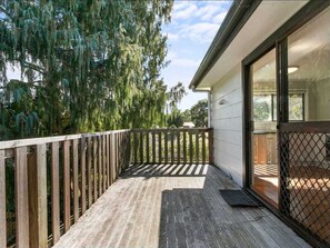 Eine Holzterrasse mit Metallgeländer, eine Glastür zu einem überdachten Bereich und Bäume im Hintergrund.