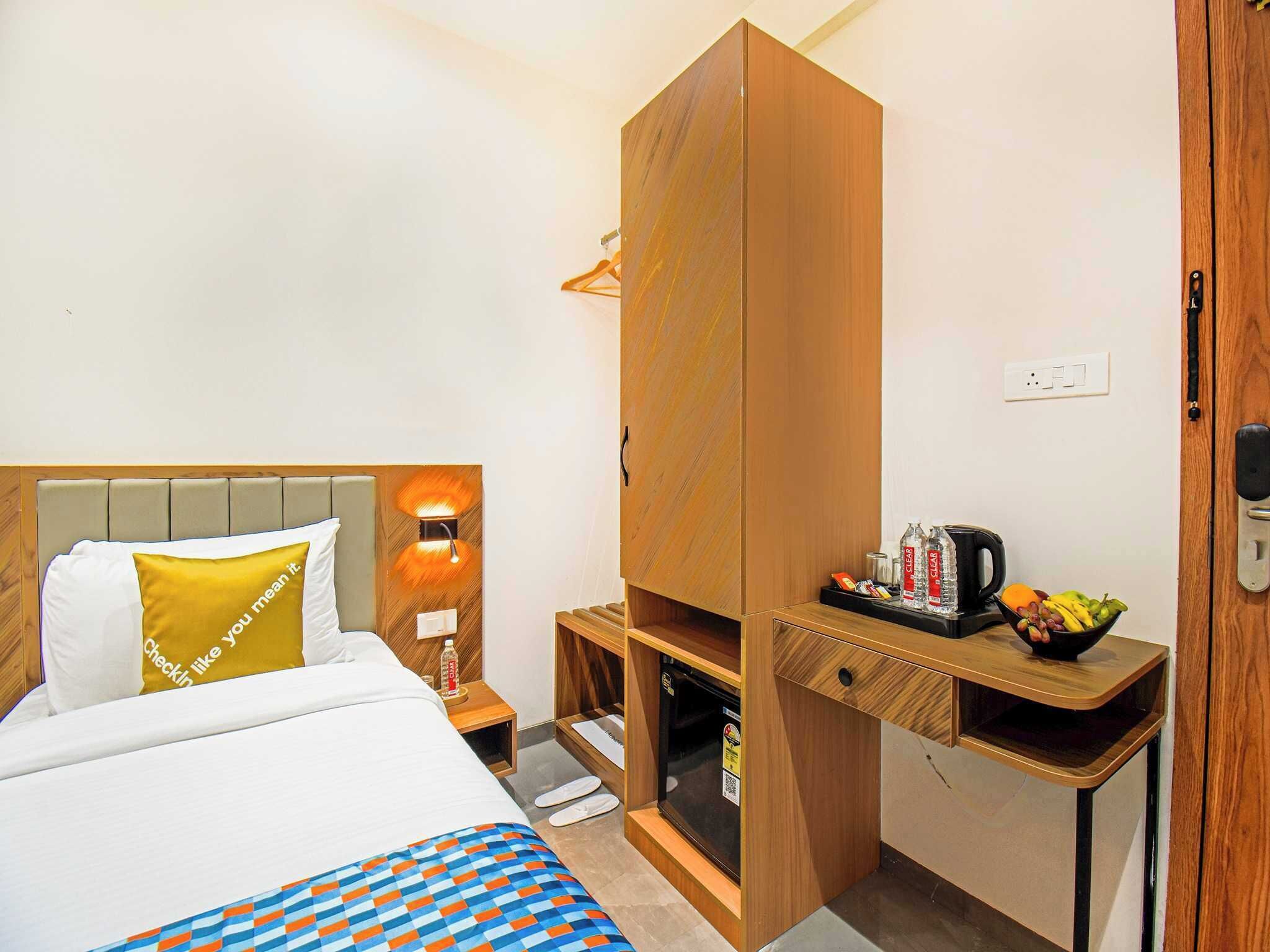 Habitación de hotel con cama, armario de madera, una mesita pequeña con una tetera y fruta, y una mesita de noche con lámpara.