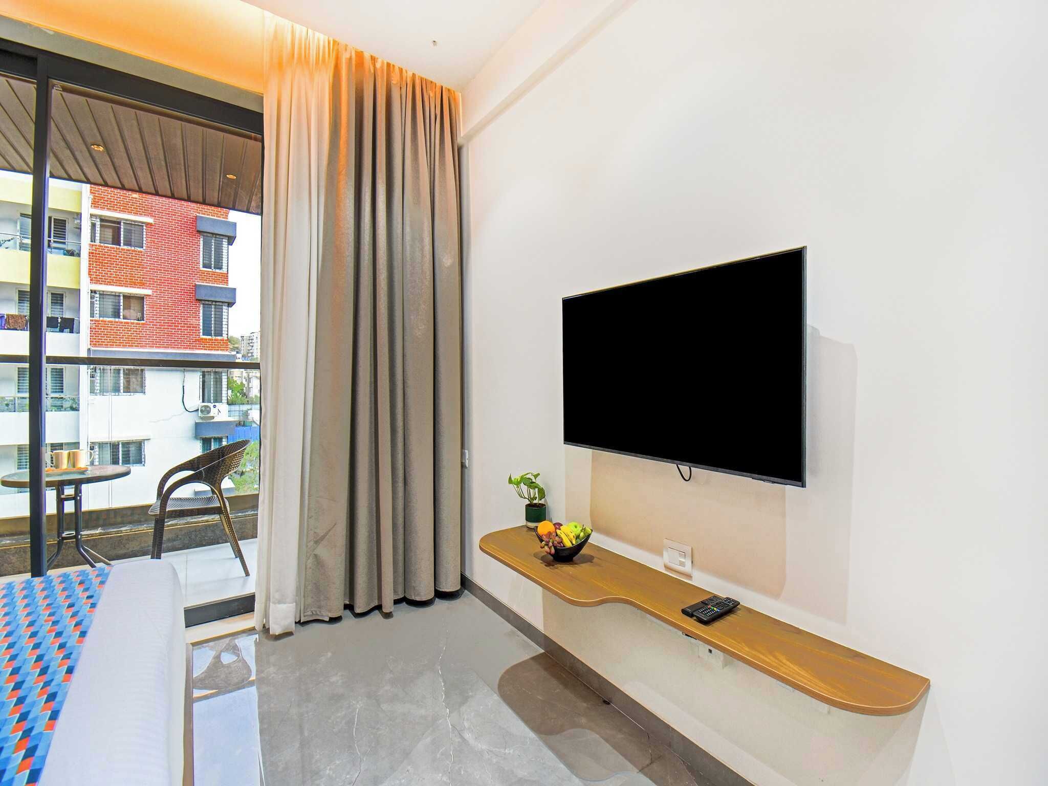 A hotel room with a large window, a flat-screen TV mounted on the wall, a small table with a chair, and a wooden bench with a bowl of fruit.