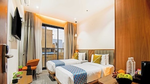 A hotel room with two beds, a desk, a chair, a TV, a balcony with curtains, and a view of buildings. - Palette Hotel and Residences Baner Pune (Pune)