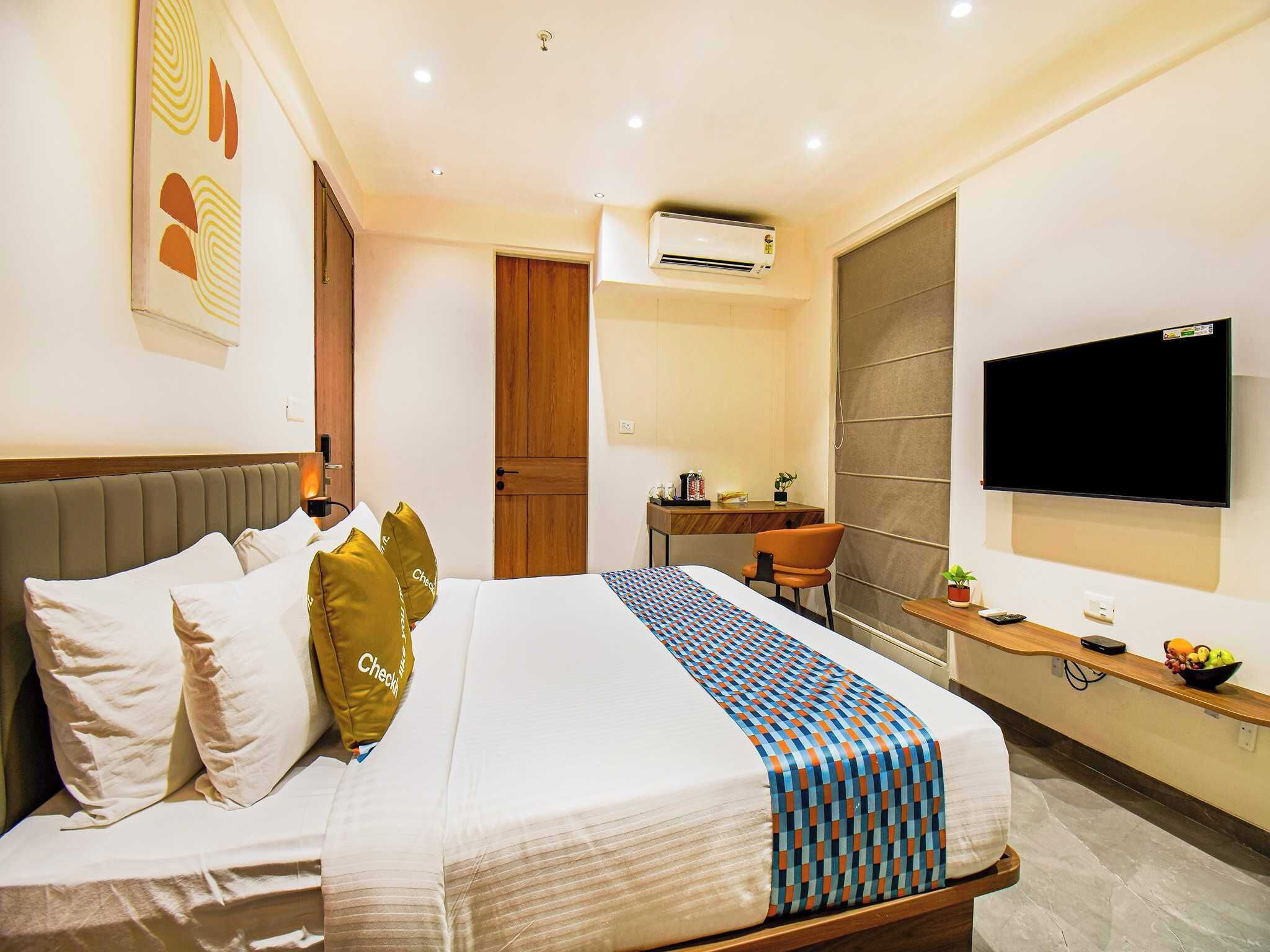 A hotel room with a bed, a desk, a chair, a TV, and an air conditioning unit.