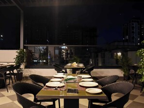 Un área de comedor al aire libre con mesas dispuestas para dos, sillas y plantas en macetas.