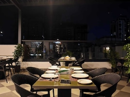 Un área de comedor al aire libre con mesas dispuestas para dos, sillas y plantas en macetas.