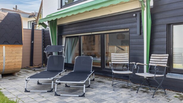 A patio with two lounge chairs and a wooden table, a wooden barrel, and a building with a green awning. - Panoramic Lake House at Steinhuder Meer – Your Exclusive Retreat by the Water (Wunstorf)