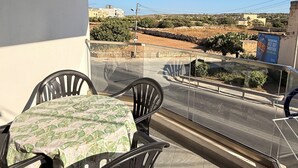 Un balcon avec une table et des chaises, donnant sur une rue bordée d’immeubles et offrant une vue sur un ciel dégagé.