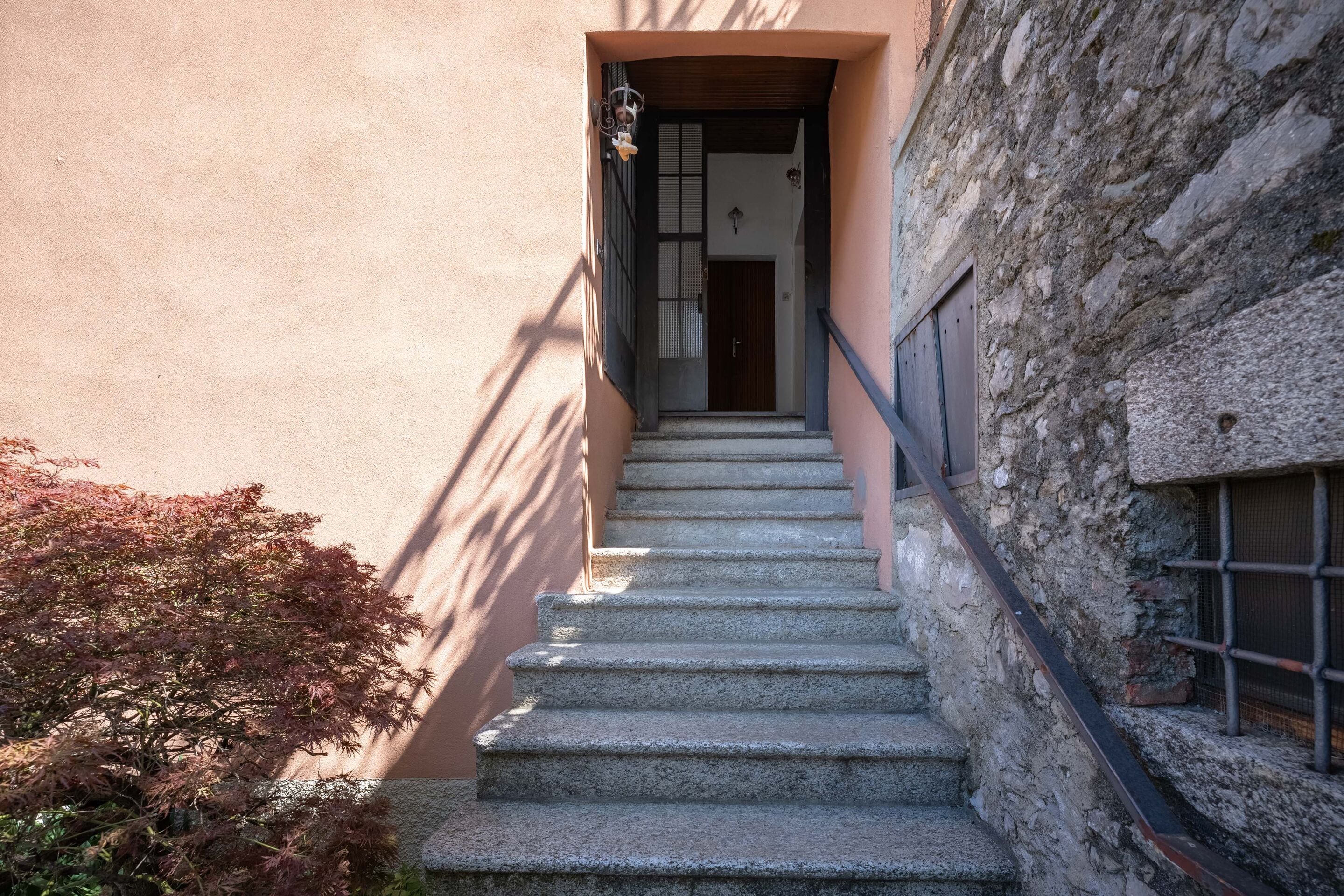 Una escalera de piedra que conduce a una puerta con reja metálica, rodeada por un edificio con pared texturizada y un pequeño arbusto.