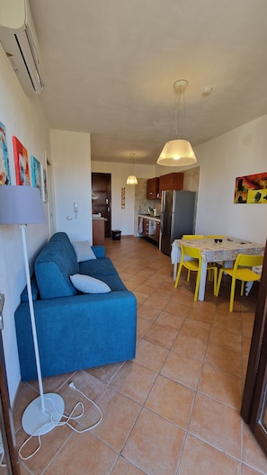 A living room with a sofa, a dining table with yellow chairs, a kitchen in the background, and a ceiling fan. - Apartment 200 meters from the beach with free parking (San Vito Lo Capo)