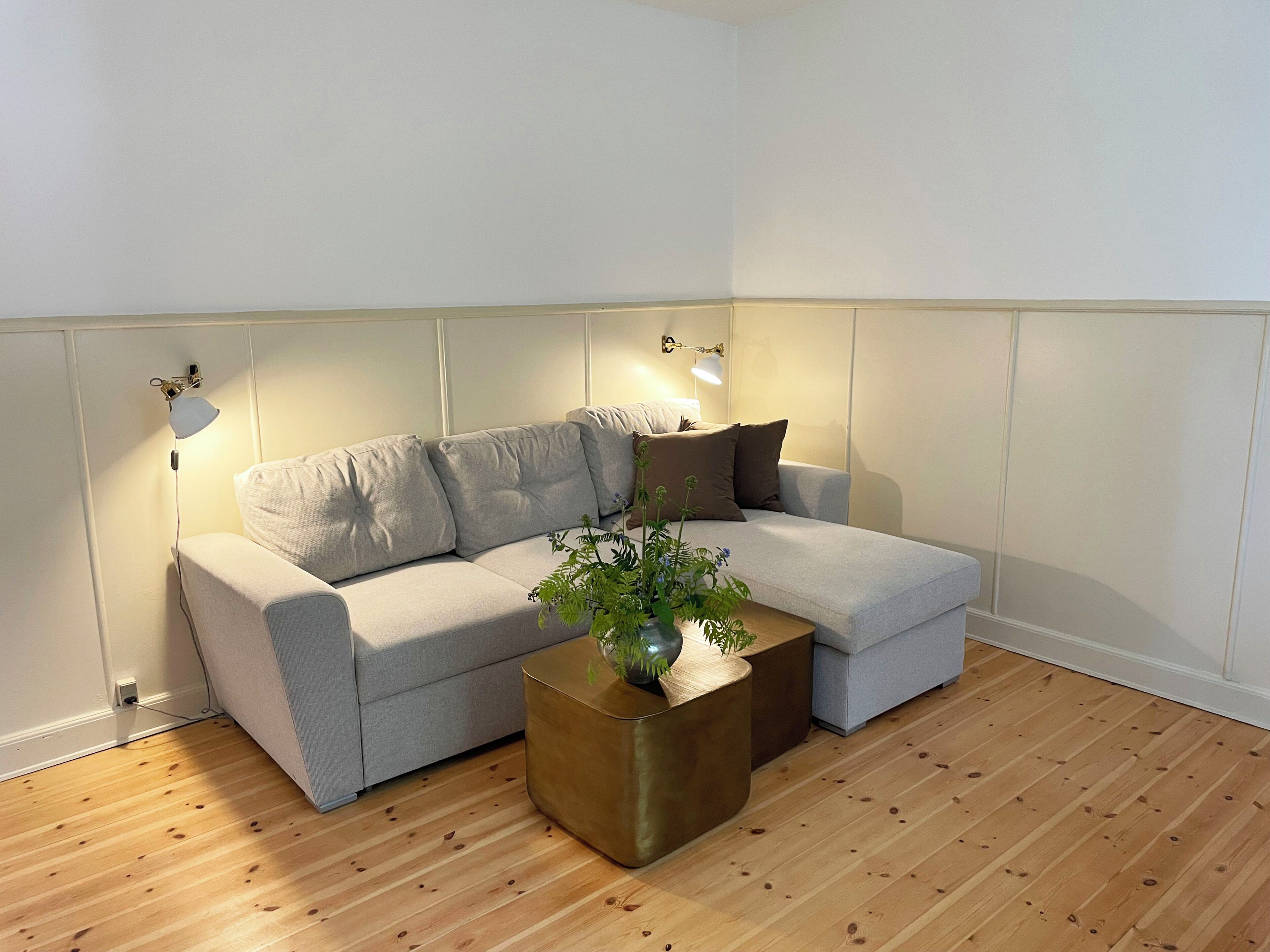 A corner living room with a grey sofa, a round wooden coffee table, and two wall-mounted lamps.
