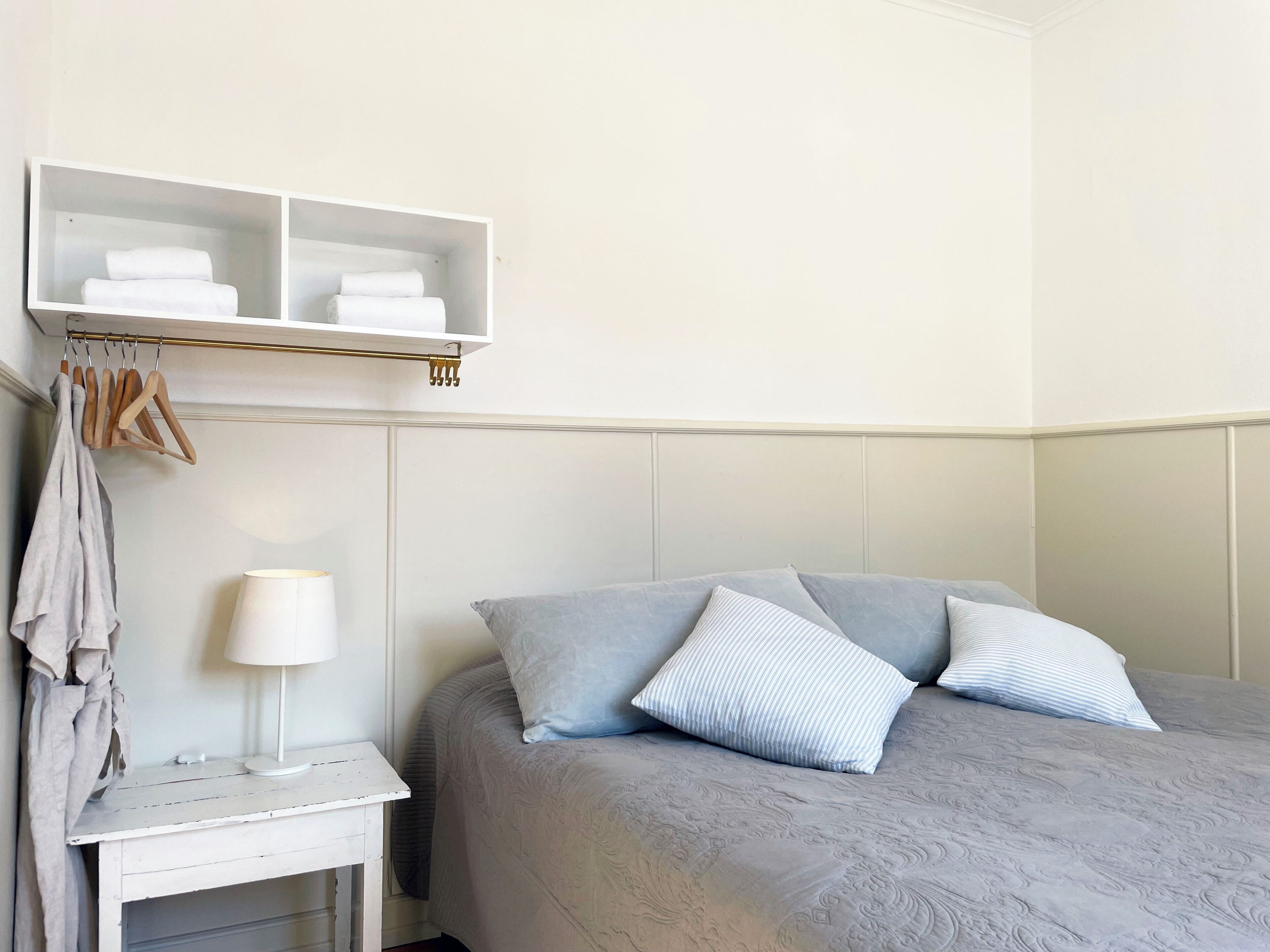 A neatly arranged hotel room with a bed, a bedside table with a lamp, a hanging clothes rack, and a shelf with folded towels.