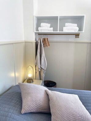Habitación de hotel ordenada con cama, almohadas, lámpara de noche, perchero con toallas y estante con toallas dobladas.