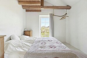 Ein Schlafzimmer mit einem Bett, einem Holz-Kopfteil, einem Nachttisch, einem Fenster mit Vorhängen und Blick ins Freie.