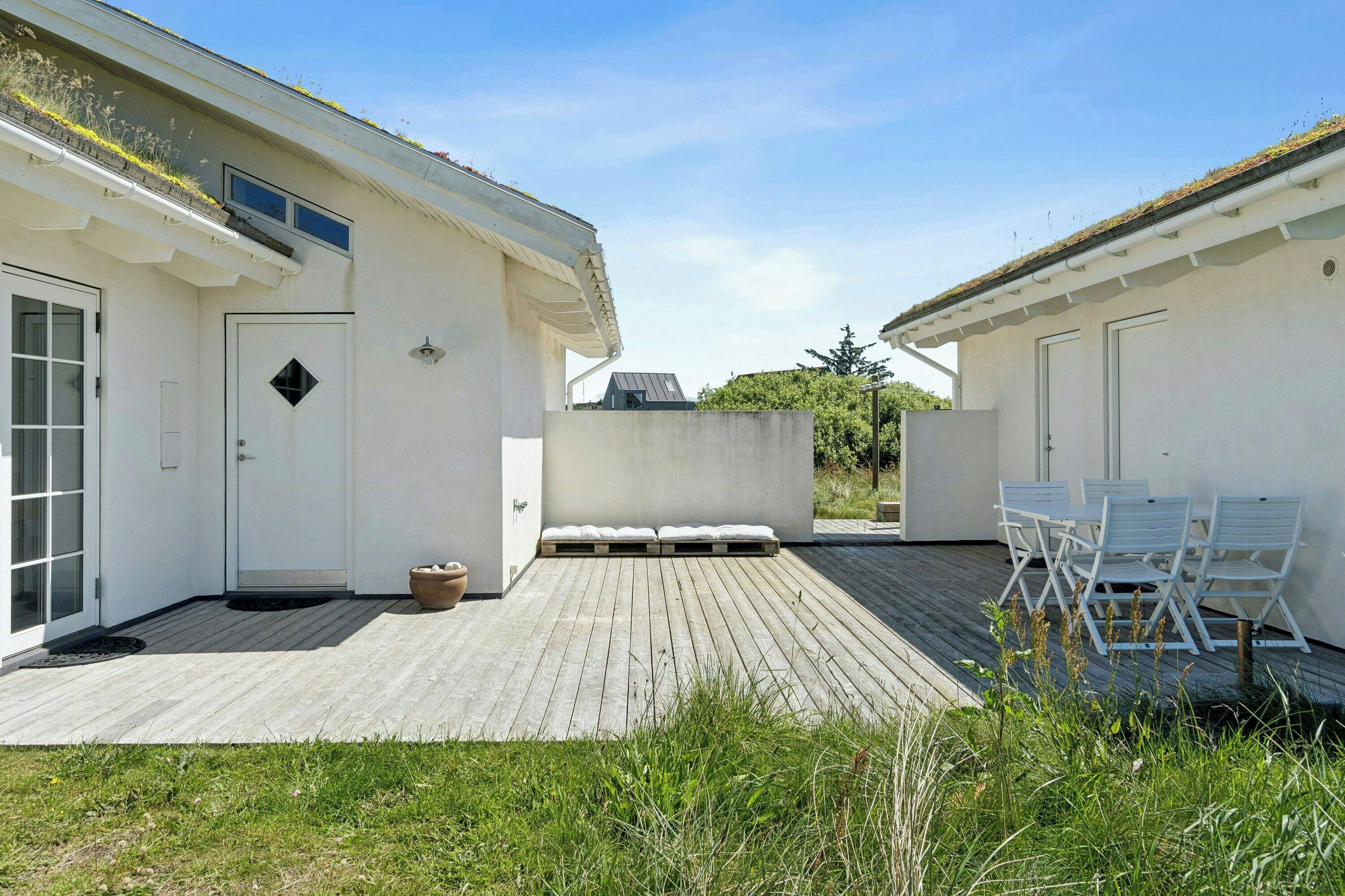 A modern building with a flat roof, a small outdoor seating area with white chairs, and a wooden deck.