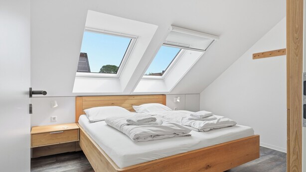 Ein Schlafzimmer mit schrägen Wänden, Dachfenstern, einem Bettgestell aus Holz und einem Nachttisch.
