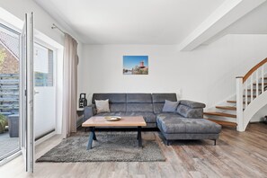 Ein modernes Wohnzimmer mit einem grauen Ecksofa, einem Holztisch und einem großen Fenster mit Blick auf eine Ziegelwand und einen roten Leuchtturm.