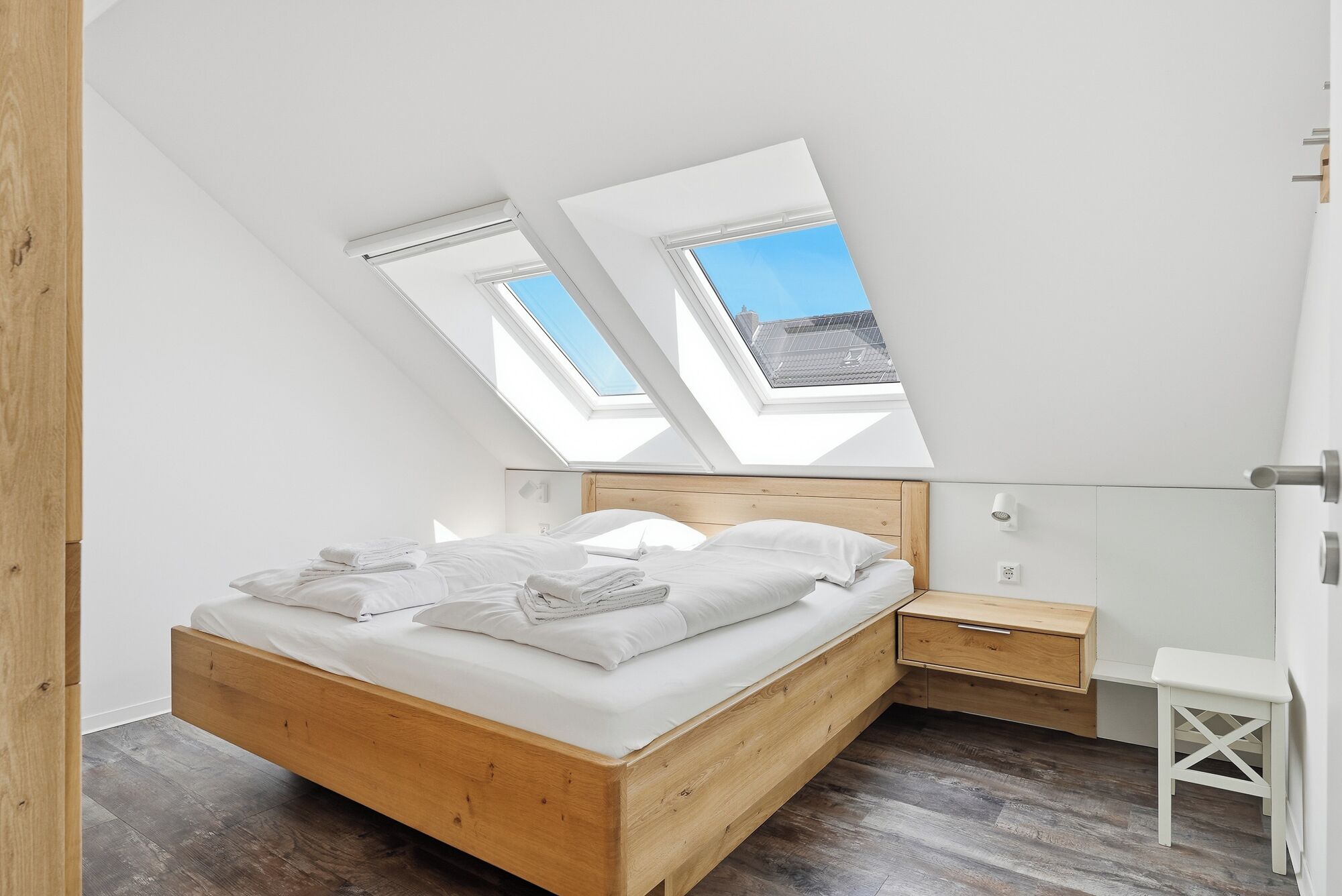 Un dormitorio con tragaluz, una cama de madera, una mesita de noche y una pequeña silla blanca.