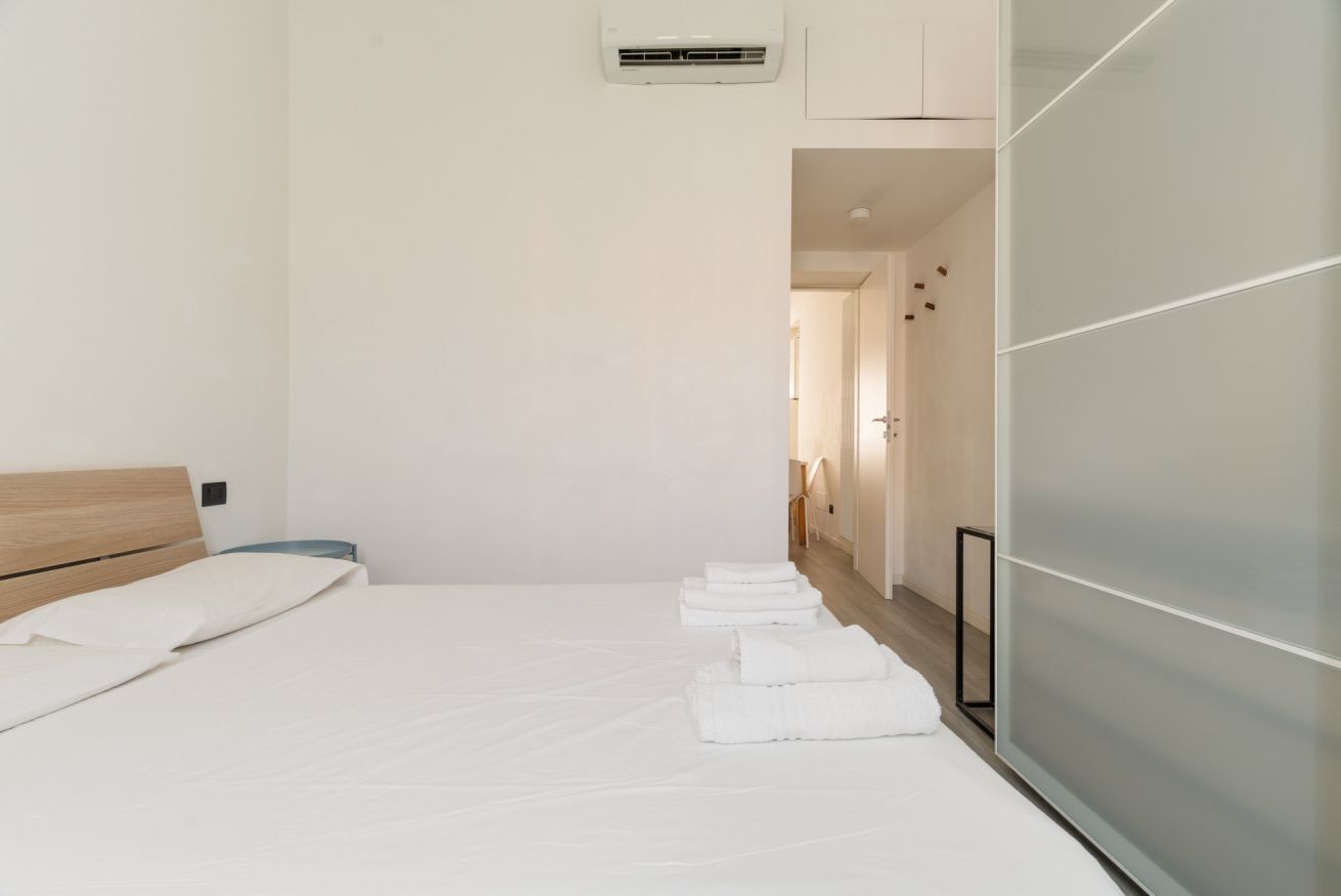 A modern hotel room with a wooden headboard, a bed with white linens, a wardrobe with frosted glass doors, and a hallway leading to another room.