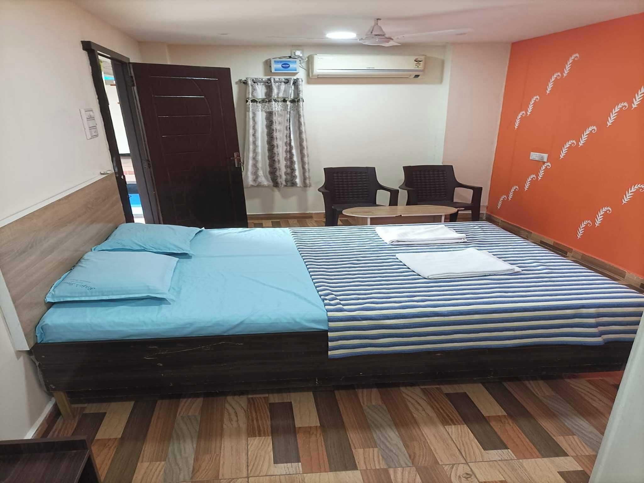 A room with a bed, two chairs, a small table, an air conditioning unit, and a door.