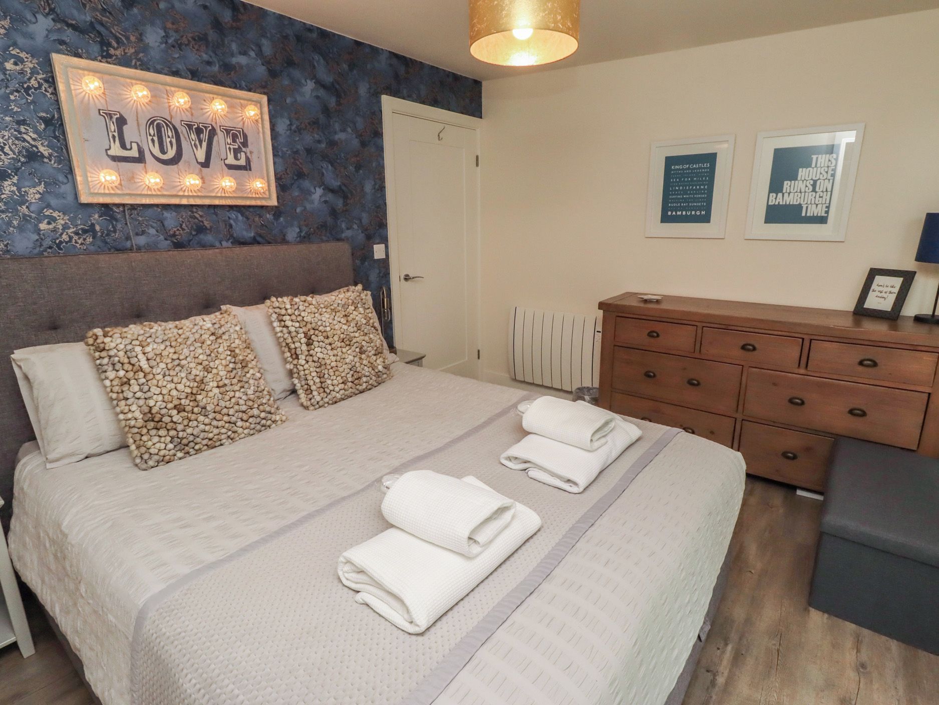 Una cama bien hecha con almohadas, una mesita de noche, un aparador y una pared con un letrero que dice 'LOVE' y cuadros enmarcados.