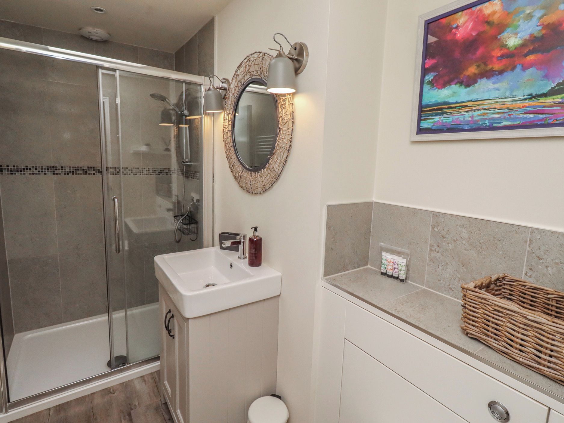 A bathroom with a glass shower enclosure, a white sink, a mirror with a decorative frame, and a painting on the wall.