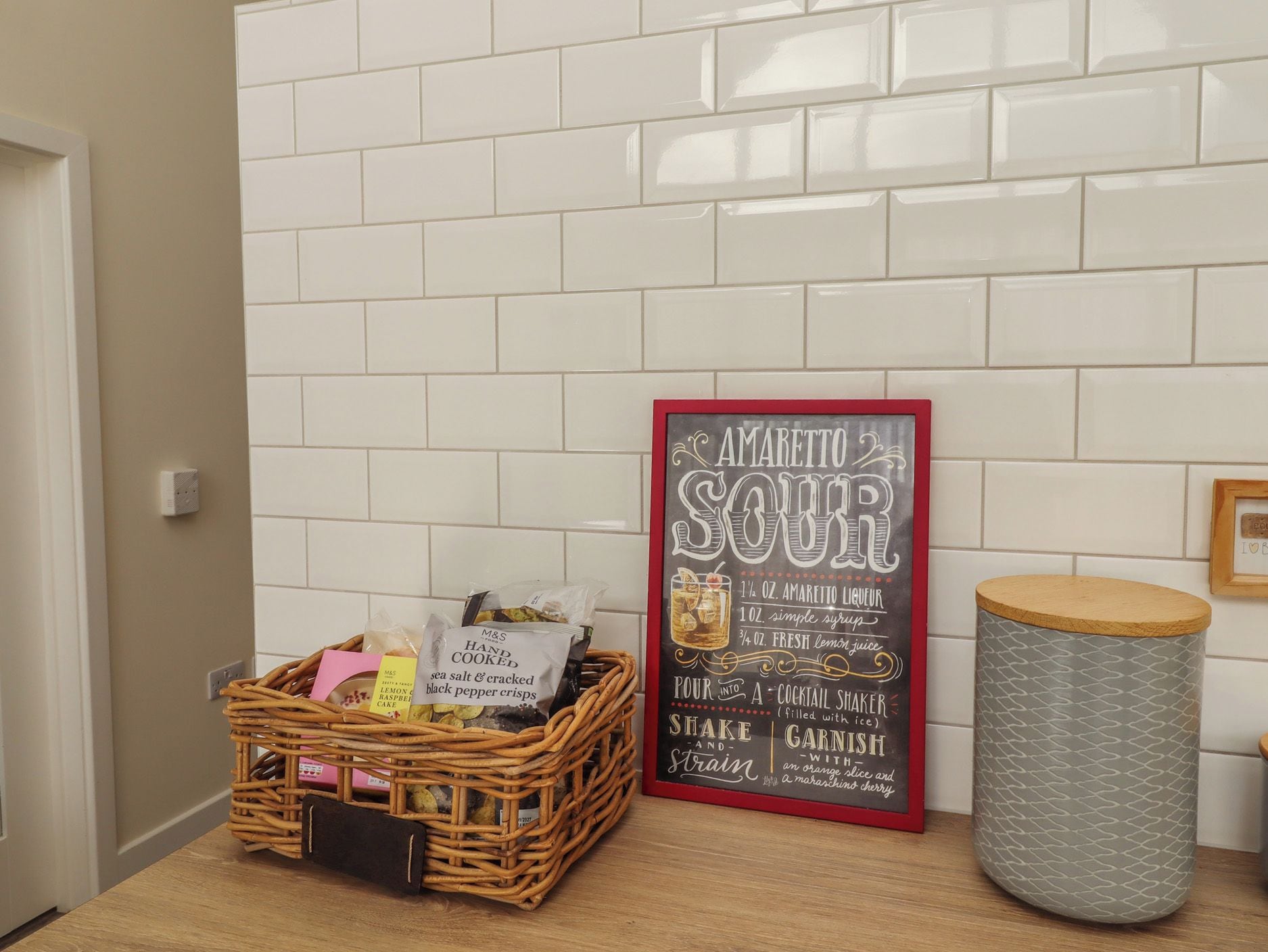 Una zona de cocina con salpicadero blanco de azulejos, una cesta de mimbre con alimentos, un cartel con recetas enmarcado y un cubo de basura.