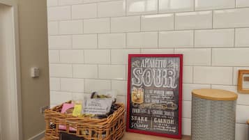 Una zona de cocina con salpicadero blanco de azulejos, una cesta de mimbre con alimentos, un cartel con recetas enmarcado y un cubo de basura.