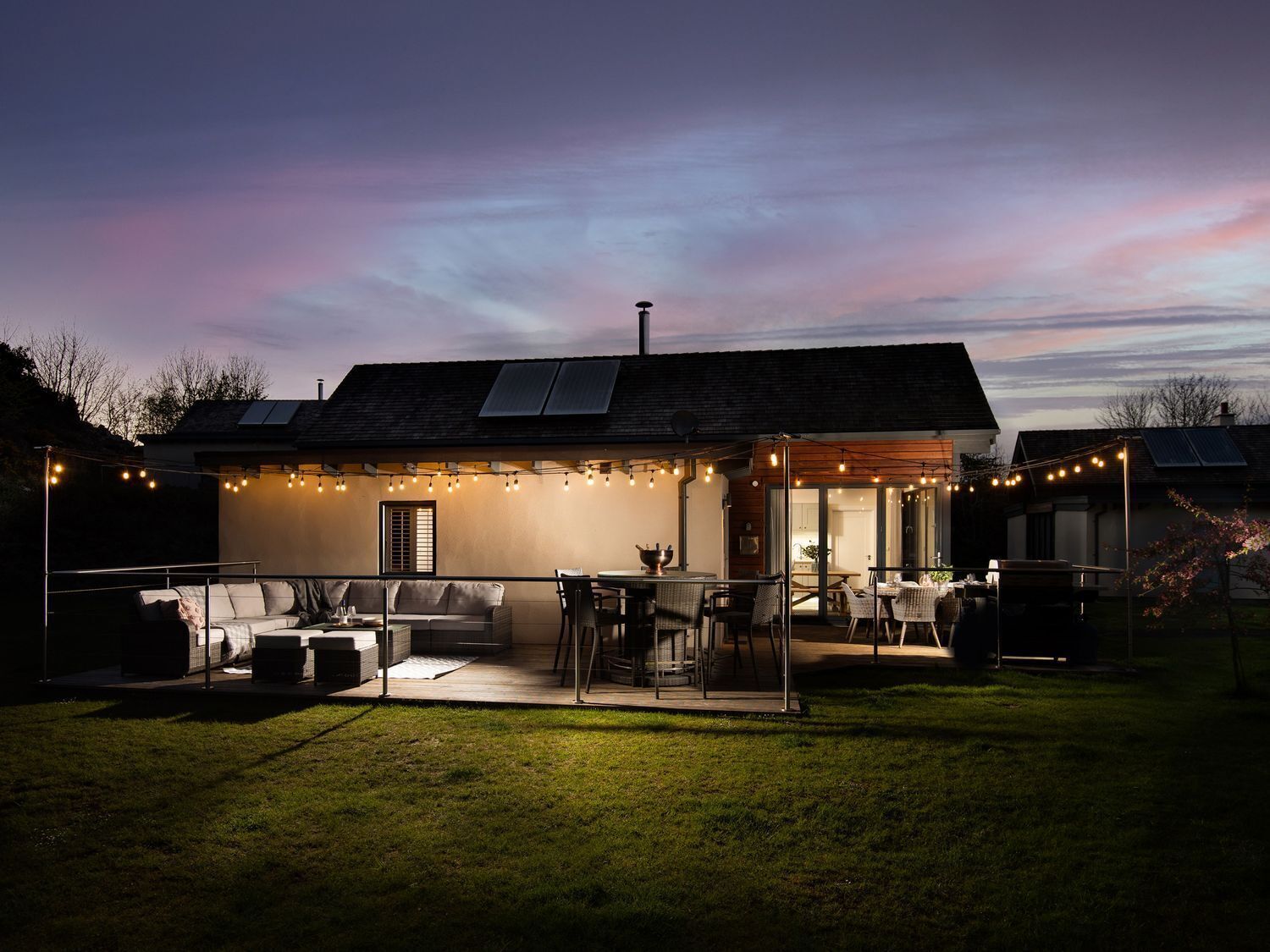 Une maison moderne dotée d’un patio, d’un espace salon extérieur et de guirlandes lumineuses.