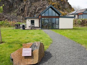 A modern lodge with a gravel path leading to the entrance, a stone with a sign, and a mountainous backdrop. - Summerset Lodge (Bamburgh)