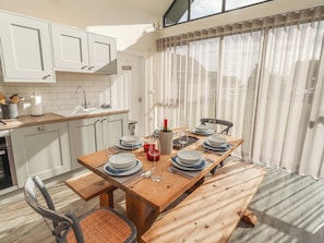 A modern kitchen with white cabinets, a wooden dining table set for six, and a large window with sheer curtains.