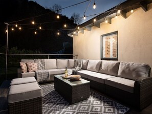 An outdoor seating area with a sofa, ottoman, and table, string lights, and a window with blinds.
