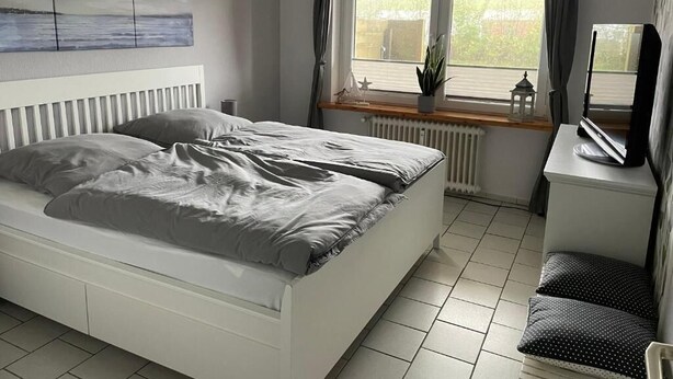 Makuuhuoneessa on sänky, työpöytä, tuoli, patteri, ikkuna, jossa on verhot, ja huonekasvi.