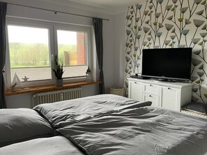 A bedroom with a bed, a TV on a cabinet, a window with curtains, and a radiator. - Stylish and authentic vacation rental in Friedrichskoog – Perfectly located (Friedrichskoog)