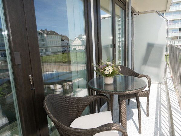 Een balkon met rieten stoelen en een glazen tafel, een bloevas en uitzicht op gebouwen door glazen deuren.