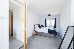 A bedroom with a bed, a window with curtains, a bedside table, and a door leading to another room. - Stylish 3-Bed Home - 10 Min Gatwick - WiFi & Parking (West Sussex)
