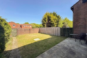 Un jardin avec une pelouse, un mur en briques, une clôture en bois et une terrasse aménagée avec du mobilier d’extérieur.