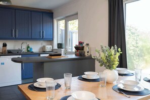 A modern kitchen with blue cabinets, a dining area with a wooden table set for four, and a large window providing natural light. - Stylish 3-Bed Home - 10 Min Gatwick - WiFi & Parking (West Sussex)