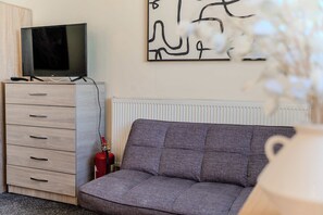 Ein Wohnzimmer mit grauem Sofa, einer Holzkassette mit Schubladen, einem Fernseher darauf und einer Heizung.