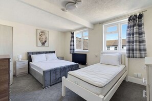 Ein Schlafzimmer mit einem Bett, einem zusätzlichen Bett, einem Schminktisch, einem Nachttisch, einem Heizkörper und einem Fenster mit Vorhängen.