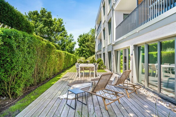 Une terrasse extérieure moderne, dotée d’une table et de chaises, entourée d’une haie bien entretenue et d’un bâtiment aux grandes fenêtres.