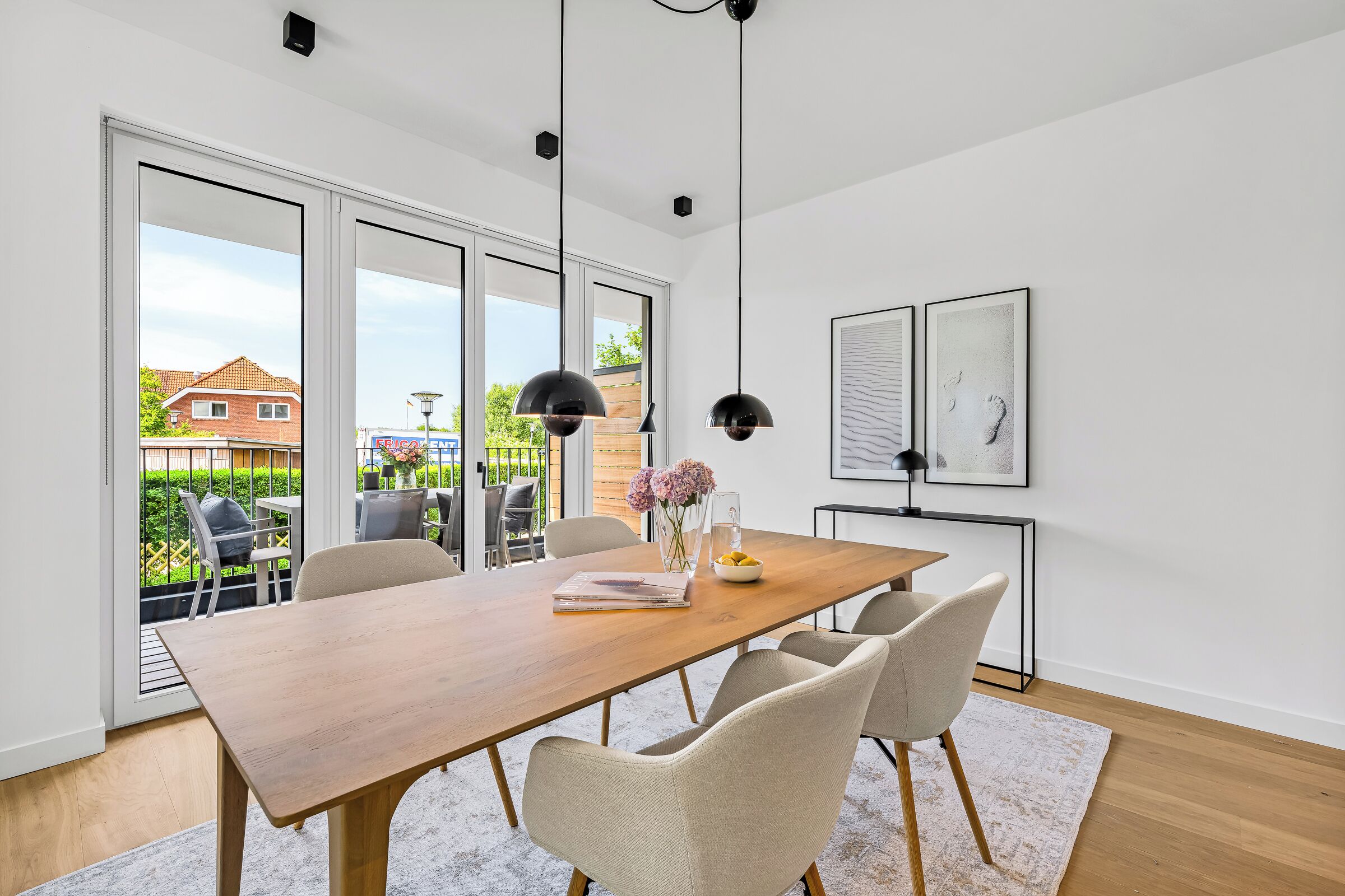 Une salle à manger moderne avec une table en bois, quatre chaises, des luminaires suspendus et une vue sur une terrasse extérieure, accessible par de grandes portes coulissantes.