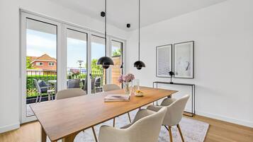 Une salle à manger moderne avec une table en bois, quatre chaises, des luminaires suspendus et une vue sur une terrasse extérieure, accessible par de grandes portes coulissantes.